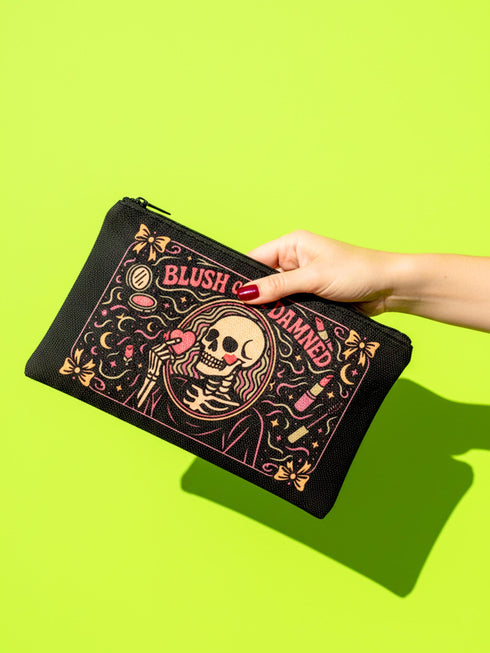 A rectangular gothic makeup bag made of black polyester with a visible woven linen-like crosshatch texture. The central illustration features a smiling human skull with flowing hair applying a pink heart-shaped blush to its cheek. Above the skull, the text BLUSH OF THE DAMNED appears. The design is framed by illustrations of lipstick tubes, mascara wands, bows. The bag includes a black zipper along the top edge and a removable black nylon wrist strap attached to the metal zipper pull.