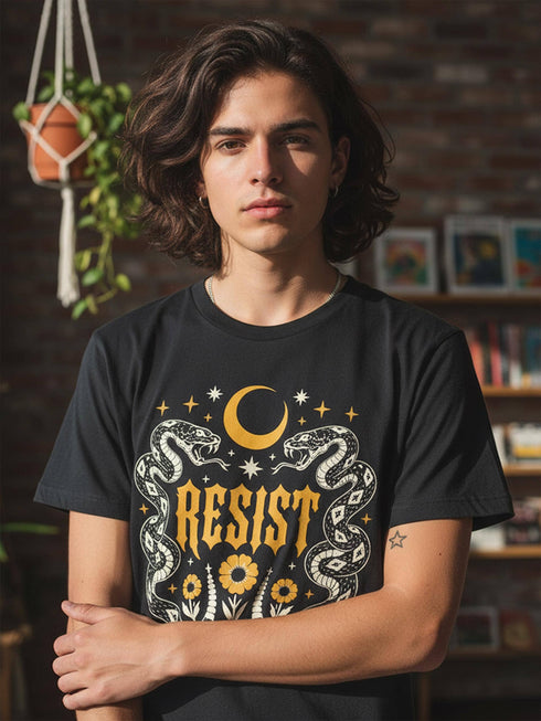 A unisex short sleeve graphic t shirt in dark charcoal black featuring a highly detailed symmetrical medallion print on the chest. In the center, the word RESIST is printed in a bold yellow gothic blackletter font. Two coiled white snakes with fine black scale details frame the text on the left and right. Above the text, a yellow crescent moon is flanked by white and yellow starbursts. Beneath, three yellow marigold flowers sit atop white stems and leafy foliage.