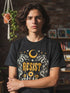 A unisex short sleeve graphic t shirt in dark charcoal black featuring a highly detailed symmetrical medallion print on the chest. In the center, the word RESIST is printed in a bold yellow gothic blackletter font. Two coiled white snakes with fine black scale details frame the text on the left and right. Above the text, a yellow crescent moon is flanked by white and yellow starbursts. Beneath, three yellow marigold flowers sit atop white stems and leafy foliage.