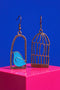 two earrings, one with a bird inside a cage and the other with a bird outside the cage, set against a vibrant blue background.