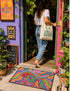 A rectangular doormat featuring a vibrant 2D flat print designed to look like textured yarn embroidery. The central graphic showcases a bright green alien with large black eyes, wearing a pink neck bandana and a highly decorated red, pink, and blue sombrero cowboy hat. The alien has one green hand raised in a friendly wave. The background is a psychedelic pattern of wavy, flowing rainbow stripes in yellow, orange, red, pink, blue, and green. The mat has a sleek black outer edge trim.
