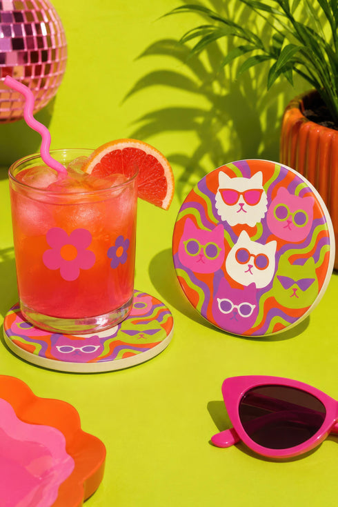 A close-up photograph of a glossy round ceramic coaster featuring a retro 70s psychedelic design. The artwork displays a cluster of six stylized cat face illustrations, each wearing a different pair of sunglasses including retro round and cat-eye shapes. The cats are colored in solid shades of white, hot pink, purple, and lime green. They are set against a vibrant background of wavy, swirling lines in bright orange, red, and purple. The top surface has a highly reflective clear glaze.