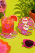 A close-up photograph of a glossy round ceramic coaster featuring a retro 70s psychedelic design. The artwork displays a cluster of six stylized cat face illustrations, each wearing a different pair of sunglasses including retro round and cat-eye shapes. The cats are colored in solid shades of white, hot pink, purple, and lime green. They are set against a vibrant background of wavy, swirling lines in bright orange, red, and purple. The top surface has a highly reflective clear glaze.