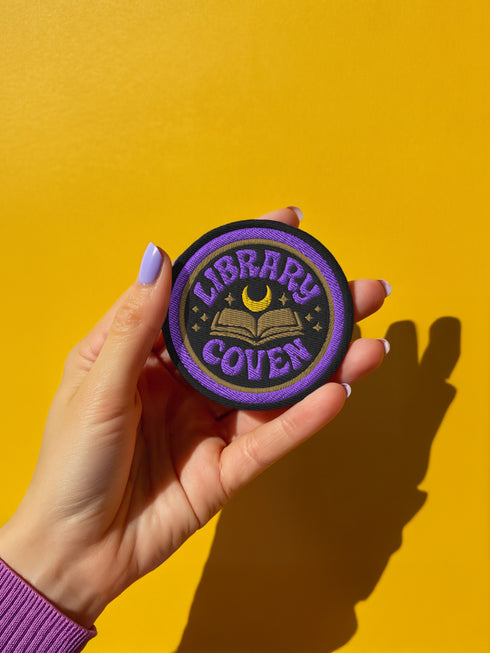 3-inch round embroidered iron-on patch in a cotton–polyester blend; black base with purple ring and gold accent ring; open book, gold crescent moon, tiny stars; curved purple text reads “LIBRARY COVEN”; merrowed edge. Witchy book lover patch for librarians, dark academia, and BookTok covens; makes a great gift for readers, witches, and spooky library pals.