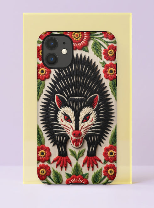 Dual layer matte phone case with a 2D print that mimics thick crewel embroidery. An angry black possum with a white face, bared sharp teeth, and bright red eyes and claws sits at the center on a cream background, framed by a symmetrical border of red folk art daisies with yellow centers and green leaves. Smooth printed surface with no raised threads, open ports, and a precise camera cutout. Goblincore, creepy cute, maximalist phone accessory.