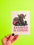 A 5x7 inch Krampus Christmas card shown in two paper finishes. The front features a stylized illustration of the folklore demon Krampus with green skin, black horns, and a pink Santa hat. He carries a large pink sack filled with three skulls over his shoulder. Below him, the words "KRAMPUS IS COMING" are printed in bold pink text. The inside of the card is blank on the left and has the printed text "Hope you've been beautifully bad this year." on the right.