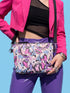 Small vegan leather crossbody purse with a real pebble grain texture and adjustable black strap. The flat printed artwork features a scattered all over pattern of pink skulls, purple and pastel blue winged bats, pink fanged mouths, and loose eyeballs set against a cream colored background. The bag is shown with black and silver hardware including a black top zipper, black zipper pull, black strap tabs, and silver metal rings and clips. The compact rectangular bag measures 8.5 by 7 by 2 inches.