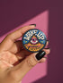 A round pinback button featuring a retro psychedelic design. The central focus is a stylized Eye of Providence in a pink triangle, surrounded by vibrant orange and yellow flames. Behind the triangle are teal and petrol blue sunburst rays. The text "WAKE UP" curves across the top in cream wavy letters, and "THE REVOLUTION" curves along the bottom in orange. The button has a glossy finish and is available in 1.75 inch or 2.25 inch sizes.