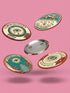 Four 2.25-inch circular metal pinback buttons on a pink textured background. The designs feature vintage tattoo-style art in rust, ochre, gold, and teal with heavy black linework. Designs include a snake Ouroboros "BREAK THE CYCLE," an umbrella eye "PROTECT THE VULNERABLE," a staircase "THE FUTURE IS OURS," and a radial eye "TRUST YOUR GAZE." The pins have a high-gloss finish. Perfect accessory for backpacks, jackets, and lanyards.