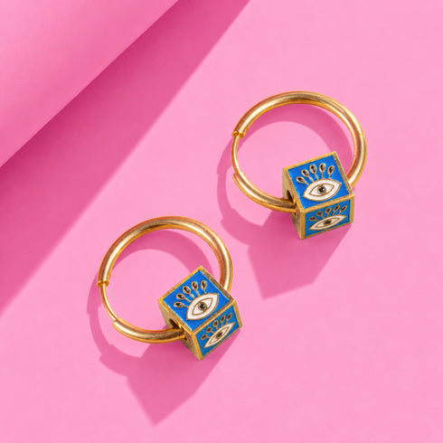 A pair of gold hoop earrings with blue enamel eye-shaped pendants, placed on a pink background.