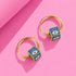A pair of gold hoop earrings with blue enamel eye-shaped pendants, placed on a pink background.