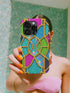 A person is holding a colorful phone case with a stained glass design, taking a selfie in front of a green tiled wall.