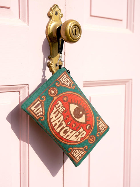 A rectangular 9x6 inch makeup bag and zipper pouch featuring a textured linen-like polyester fabric. The bag showcases a bold, maximalist print with vivid colors typical of retro 70s and pastel goth aesthetics, centering on a large mystic eye design labeled "The Watcher." It features a sturdy black zipper across the top and includes a removable black nylon wristlet strap.Perfect for use as a pencil case, cosmetic organizer, or festival wristlet.