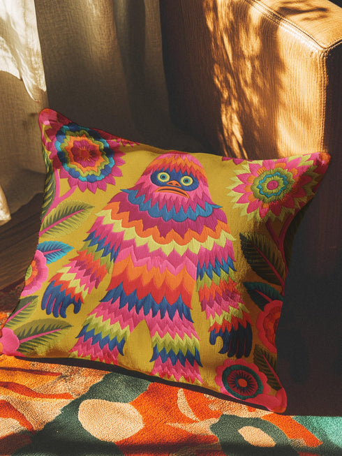 A square faux embroidery pillow featuring a brightly colored maximalist design against a mustard yellow background. In the center stands a stylized, furry Bigfoot or Sasquatch figure rendered in vibrant layers of pink, orange, blue, and lime green with a blue face and yellow eyes. The central creature is surrounded by large, detailed symmetrical pink and blue flowers and leaves. The flat printed fabric creates a highly realistic optical illusion of dimensional crewel embroidery stitches.