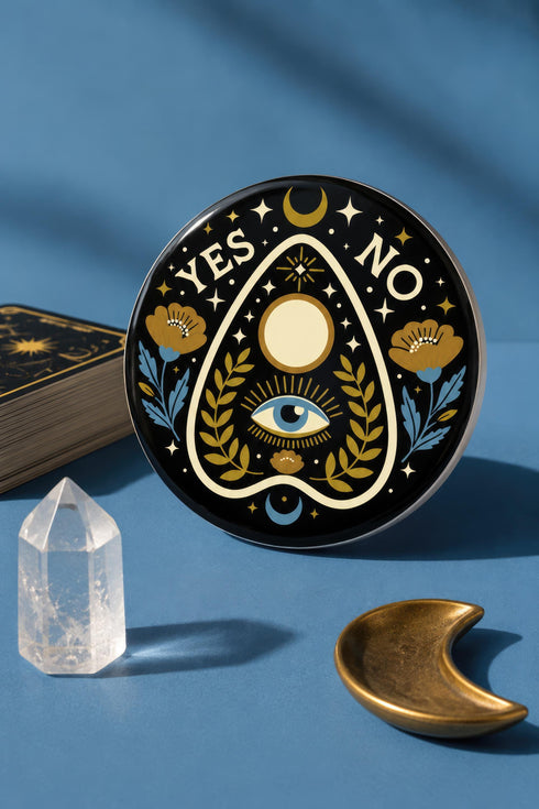 A close-up photograph of a glossy ceramic coaster featuring a Planchette occult design on a deep black background. The symmetrical graphic displays a central blue All-Seeing Eye with gold sunburst lines, topped by a white circle, and flanked by gold flowers with blue stems. At the top, the text "YES" is printed in gold on the left and "NO" on the right. Gold stars and crescent moons are scattered around the perimeter. The flat, round coaster has a highly visible high-gloss glaze.