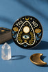 A close-up photograph of a glossy ceramic coaster featuring a Planchette occult design on a deep black background. The symmetrical graphic displays a central blue All-Seeing Eye with gold sunburst lines, topped by a white circle, and flanked by gold flowers with blue stems. At the top, the text "YES" is printed in gold on the left and "NO" on the right. Gold stars and crescent moons are scattered around the perimeter. The flat, round coaster has a highly visible high-gloss glaze.