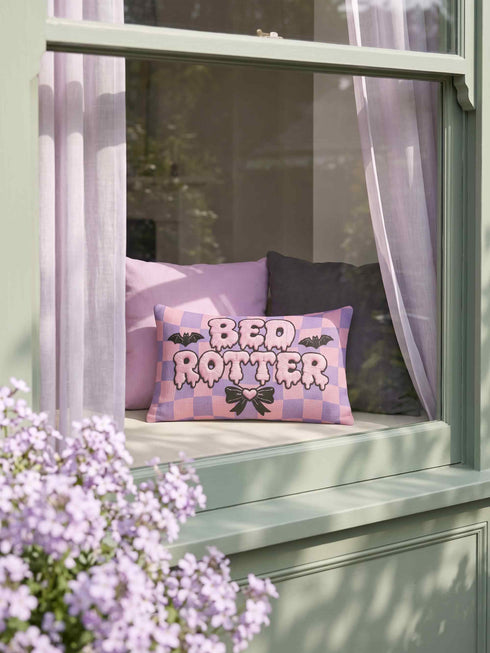 Pastel goth lumbar pillow, 20 by 12 inches, with a pink and lavender purple wavy checkerboard background. The words Bed Rotter appear in dripping pink bubble letters with a faux embroidery thread texture and black outlines. Two black bats with pink stitch details flank the text, and a black bow with a pink heart center sits below. Double sided flat print that mimics raised embroidery on linen feel polyester with a hidden zipper, insert included.