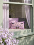 Pastel goth lumbar pillow, 20 by 12 inches, with a pink and lavender purple wavy checkerboard background. The words Bed Rotter appear in dripping pink bubble letters with a faux embroidery thread texture and black outlines. Two black bats with pink stitch details flank the text, and a black bow with a pink heart center sits below. Double sided flat print that mimics raised embroidery on linen feel polyester with a hidden zipper, insert included.