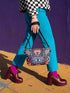 Small vegan leather crossbody purse with a real pebble grain texture and adjustable black strap. The flat printed artwork features a central blue sunburst evil eye motif below a coral pink sacred heart eye, surrounded by pink crescent moons, multicolored stars, flowing teal botanical vines, and wavy lavender and black checkerboard distortion banners. Set against a dark background with silver metal hardware including a top zipper, zipper pull, and strap clips. Measures 8.5 by 7 by 2 inches.
