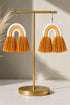 A pair of gold-colored earrings with orange tassels hanging from them, displayed on a gold stand.