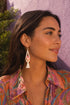A woman with long dark hair wearing a colorful patterned shirt and a white tassel earring is smiling and looking to the side.