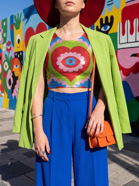 A sleeveless scoop neck crop top featuring a bold all-over printed design. The central graphic is a large bright red heart enclosing a scalloped pink flower shape and a blue evil eye motif in the middle. The background surrounds the heart with mirrored concentric wavy arches and scalloped borders in vibrant shades of lime green, pink, blue, and red. The fitted garment has a smooth athletic texture. Available in a standard matte performance knit or a sparkly silver glitter foil finish.