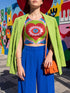 A sleeveless scoop neck crop top featuring a bold all-over printed design. The central graphic is a large bright red heart enclosing a scalloped pink flower shape and a blue evil eye motif in the middle. The background surrounds the heart with mirrored concentric wavy arches and scalloped borders in vibrant shades of lime green, pink, blue, and red. The fitted garment has a smooth athletic texture. Available in a standard matte performance knit or a sparkly silver glitter foil finish.