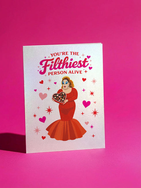 A 5x7 inch pearlescent silver greeting card featuring a flat vector illustration of the drag queen Divine in a red mermaid gown holding a heart-shaped box of chocolates. The text above reads "You're The Filthiest Person Alive" in bold red script with pink sparkles. The card has a shimmering finish that reflects light. Inside text reads "...and I love it." The back features the Honey Dazed logo. Includes a matching envelope.