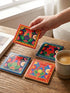 A set of four square coasters featuring a glossy glazed finish and vibrant insect illustrations. Each coaster measures approximately four by four inches. The designs showcase symmetrical beetles and spiders in bold red, blue, and orange colors, framed by intricate floral and geometric borders in a maximalist folk art style. Available in ceramic with cork backing or hardboard MDF. The edges are smooth. The designs are vivid and highly saturated, designed to provide grip and protect furniture.