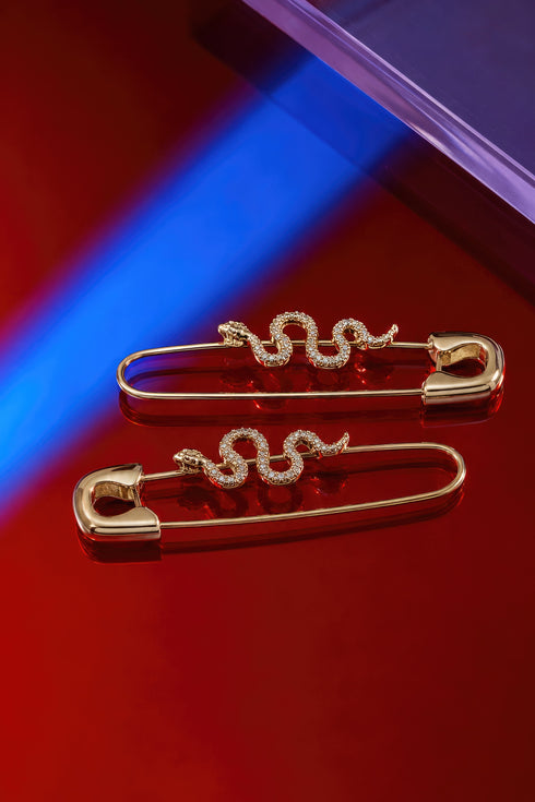 two gold-colored safety pins with snake-shaped designs on them, placed on a red surface.