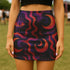 High-waisted mini skirt featuring a surreal wavy pattern with all-seeing eye and crescent moon motifs in magenta, plum, and burnt orange. Available in metallic or performance knit finishes. Sizes XS to XL.