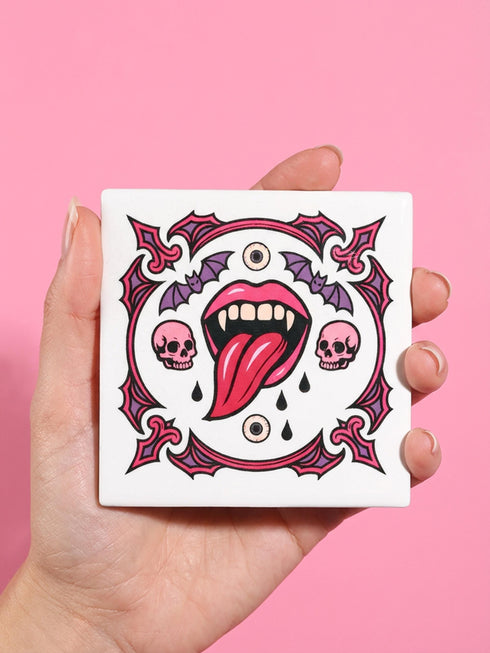 White porcelain 11oz mug and matching 4x4 inch square ceramic coaster set featuring a vibrant, maximalist full-wrap pattern. The design illustrates glossy pink vampire lips with white fangs and red tongues, interspersed with floating eyeballs, purple bats, and small pink skulls against a white background. The mug features a glossy finish and C-handle. The coaster has a matching glazed ceramic top and a brown cork backing. High-contrast pop-art style with pink, purple, and black details.