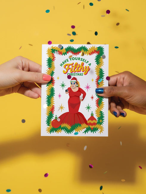 A 5x7 pearlescent holiday card by HoneyDazed. The card illustration, inspired by John Waters, features the drag queen Divine in a red dress with a Santa hat. She points a gun, withtext reads "Have Yourself a Filthy Christmas" in campy font. The card is bordered by garland. Iinside of the card reads: "Season's Sleazings" in red and green text. This kitschy greeting card is a perfect piece of cult film memorabilia and a funny adult humor gift for fans of queer art.