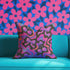 Throw pillow with a black background and a vibrant Y2K-inspired design featuring gradient cherries and shiny bows. The pattern includes pink, purple, orange, and blue psychedelic squiggles. The square pillow is displayed on a pink chair, bright blue couch, and outdoors with camping gear, emphasizing its bold, girly, and playful aesthetic. Perfect for dopamine decor, Gen Z bedrooms, or quirky home styling.