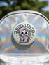 Round pinback button featuring a pastel mint green background with a cartoon grim reaper character in a lavender hood holding a protest sign. The design includes bold black outlined lettering reading “Pacifist Reaper” above the figure and small sparkle accents surrounding it. The finish is glossy and smooth, with a printed illustration style on a metal pin badge surface.
