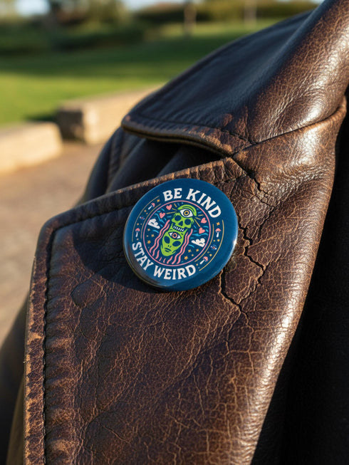A round 1.75 inch pinback button featuring a retro psychedelic design on a dark navy blue background. The text reads BE KIND STAY WEIRD in vintage cream lettering around the border. The center illustration depicts a neon green skull stacked on top of a neon green alien head. Both the skull and alien have opening third eyes on their foreheads. Pink wavy lines frame the faces. The background is decorated with small colorful mushrooms, hearts, stars, and clouds. Glossy finish.
