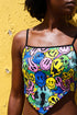 A sleeveless women's camisole top featuring a pointed handkerchief hemline often called a bandana top style. The fabric displays a vibrant all-over psychedelic print of distorting and melting smiley faces mixed with melting skulls. The color palette includes bright neon yellow, electric blue, hot pink, and lime green against a multi-colored background. The top features thin black adjustable spaghetti straps and black trim along the straight neckline. The fit is cropped and form-fitting.