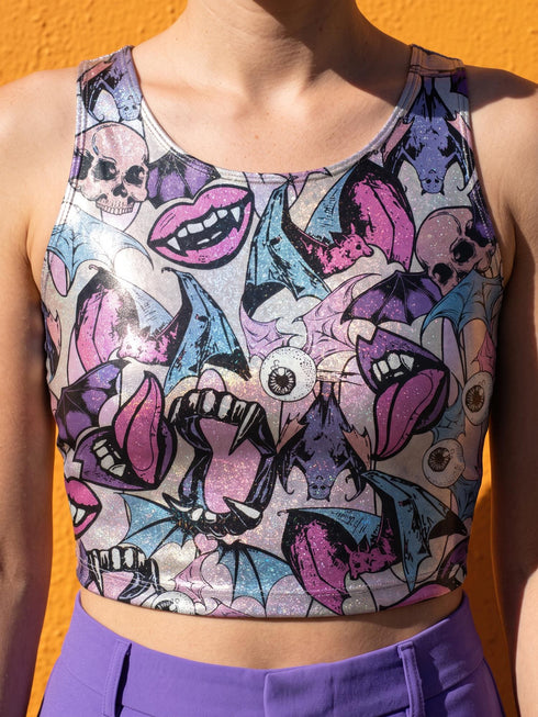A sleeveless cropped tank top made from stretchy performance knit fabric. The all-over print features a dense, scattered pattern of pastel goth and creepy cute motifs over a light background. Graphics include pink and blue bats in flight, floating eyeballs with blue irises, pink skulls, and glossy vampire lips with sharp white fangs and tongues sticking out. The neckline is a high scoop and the armholes are curved. The fabric comes in either a matte finish or a sparkly silver glitter foil.