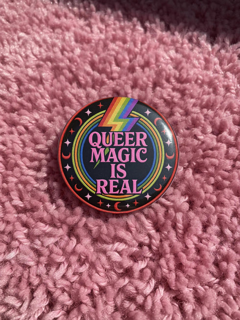 A round metal pinback button badge featuring a black background with the words QUEER MAGIC IS REAL in capital pink retro serif typography. A bright rainbow colored lightning bolt strikes from the top center, surrounded by concentric rainbow rings in red, orange, yellow, green, blue, and purple. The design is accented with small pink four-point stars and red crescent moons. The artwork features a realistic 2-D faux embroidery texture. The back has a silver metal safety pin clasp fastening.