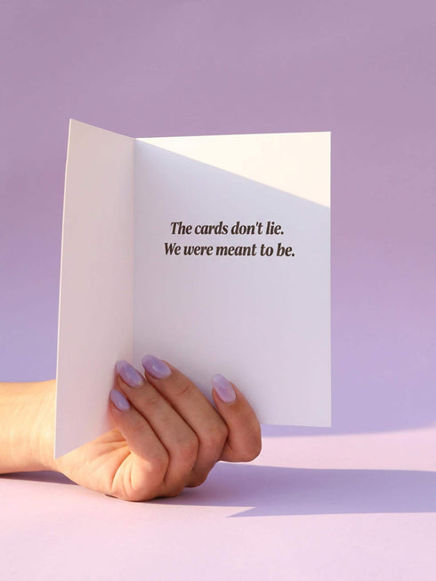 A vertical folded greeting card featuring a pastel goth tarot design titled The Devotion. The central illustration shows a pale devil imp with black wings and horns, holding a red heart in the left hand and a black dagger in the right. The imp has one star eye and one heart eye. The background is lavender purple. A black border frames the scene with teal vines, skulls, gold crescent moons, and roses. The bottom banner reads YOU + ME. The finish is shimmering pearlescent.