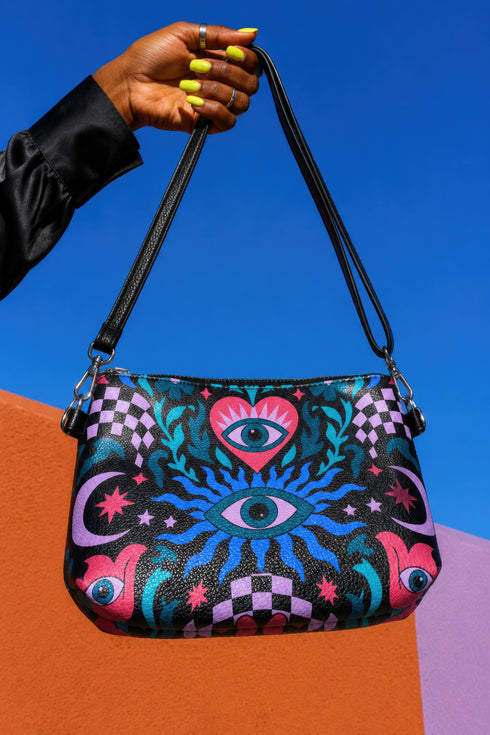 Small vegan leather crossbody purse with a real pebble grain texture and adjustable black strap. The flat printed artwork features a central blue sunburst evil eye motif below a coral pink sacred heart eye, surrounded by pink crescent moons, multicolored stars, flowing teal botanical vines, and wavy lavender and black checkerboard distortion banners. Set against a dark background with silver metal hardware including a top zipper, zipper pull, and strap clips. Measures 8.5 by 7 by 2 inches.