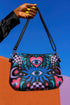 Small vegan leather crossbody purse with a real pebble grain texture and adjustable black strap. The flat printed artwork features a central blue sunburst evil eye motif below a coral pink sacred heart eye, surrounded by pink crescent moons, multicolored stars, flowing teal botanical vines, and wavy lavender and black checkerboard distortion banners. Set against a dark background with silver metal hardware including a top zipper, zipper pull, and strap clips. Measures 8.5 by 7 by 2 inches.