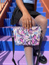Small vegan leather crossbody purse with a real pebble grain texture and adjustable black strap. The flat printed artwork features a scattered all over pattern of pink skulls, purple and pastel blue winged bats, pink fanged mouths, and loose eyeballs set against a cream colored background. The bag is shown with black and silver hardware including a black top zipper, black zipper pull, black strap tabs, and silver metal rings and clips. The compact rectangular bag measures 8.5 by 7 by 2 inches.