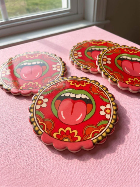 Round ceramic coaster with scalloped edges featuring a vibrant pop art design of an open mouth with green lips and a red tongue sticking out. The background is a bright red with psychedelic floral swirls in yellow orange and green. The surface is glossy glazed ceramic. The back is made of cork to prevent slipping. This maximalist decor piece fits retro 70s funky and weirdcore aesthetics. It is a bold statement piece for coffee tables. The design is colorful and eclectic dopamine decor.