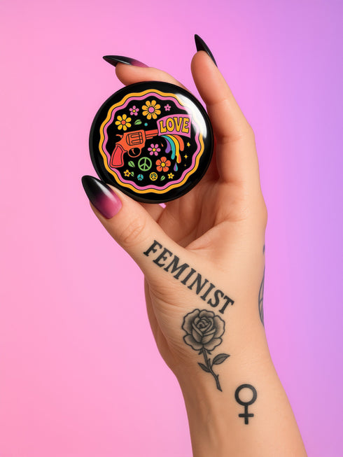 Round pinback button badge with a black background, featuring a retro illustration of a red revolver shooting out a rainbow flag that says “LOVE.” Surrounded by colorful 70s-style flowers and peace signs, outlined in wavy neon border. Glossy finish with vibrant pop colors and psychedelic design.