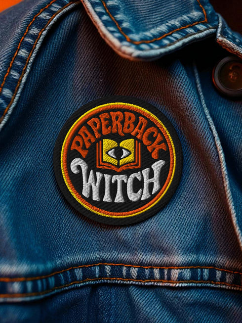 A round, black embroidered patch with a thick, textured merrow border stitched in alternating orange and yellow thread. In the center, the words "PAPERBACK WITCH" are embroidered in a retro 70s-style font. The word "PAPERBACK" is arched across the top in bold orange thread with a thin yellow outline. The word "WITCH" is across the bottom in puffy, bright white thread with a thin yellow outline. Between the words is a central icon of an open book, containing a stylized all-seeing eye.