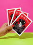 A vertical greeting card featuring a flat vector illustration of Robert Smith from The Cure against a vibrant red background. He has messy black hair, pale skin with smeared lipstick, and wears black cat ears. He is surrounded by four black cats with pink collars. White script text at the top reads "Be my Lovecat." The card is printed on shimmery pearlescent cardstock that catches the light. A high-quality alternative Valentine or anniversary card for goth music lovers and post-punk fans.
