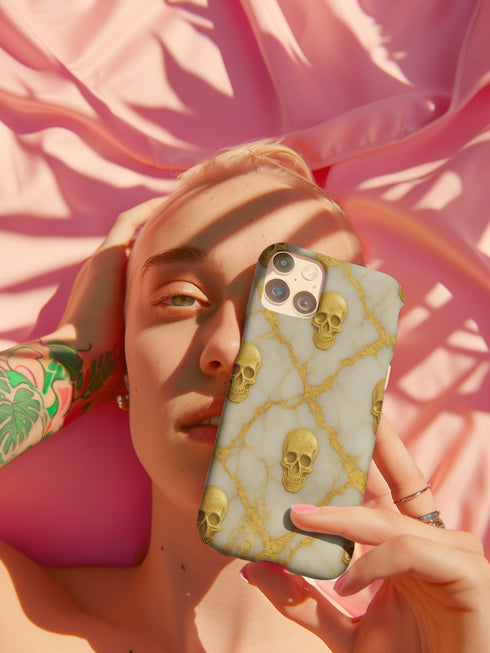 A person with tattoos is holding a phone with a skull design case up to their face, partially obscuring their face.