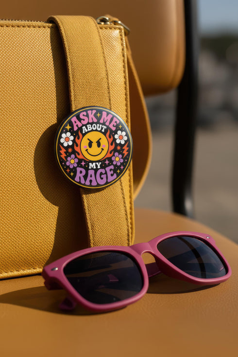A round glossy metal pinback button featuring a flat 2D print design on a solid black background. In the center is an angry yellow smiley face surrounded by orange and red flames. Above the smiley face, pink text reads ASK ME and white text reads ABOUT. Below the face, white text reads MY and large purple text reads RAGE. The design is accented with two white daisies, two purple flowers, pink and orange four-point stars, and orange lightning bolts framing the outer edges.