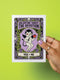 A vertical folded greeting card featuring a pastel goth tarot design titled The Devotion. The central illustration shows a pale devil imp with black wings and horns, holding a red heart in the left hand and a black dagger in the right. The imp has one star eye and one heart eye. The background is lavender purple. A black border frames the scene with teal vines, skulls, gold crescent moons, and roses. The bottom banner reads YOU + ME. The finish is shimmering pearlescent.